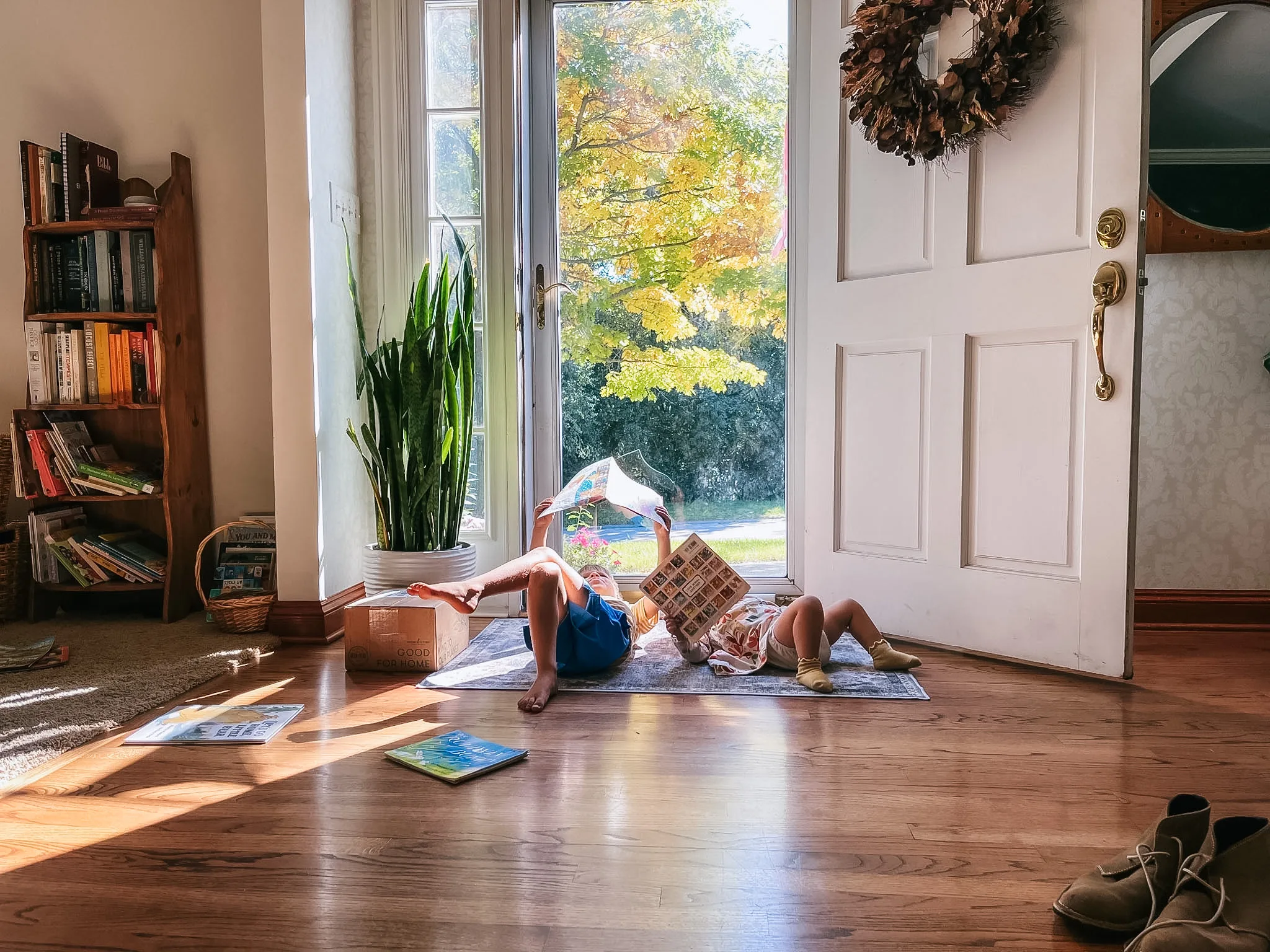 Two children lying on a rug in a sunlit entryway, reading books, with autumn trees visible through the glass door.